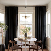 Floor-length black curtains frame a bright dining room window behind a round white table with wooden chairs, stacked dishes, and a vase of leafy branches