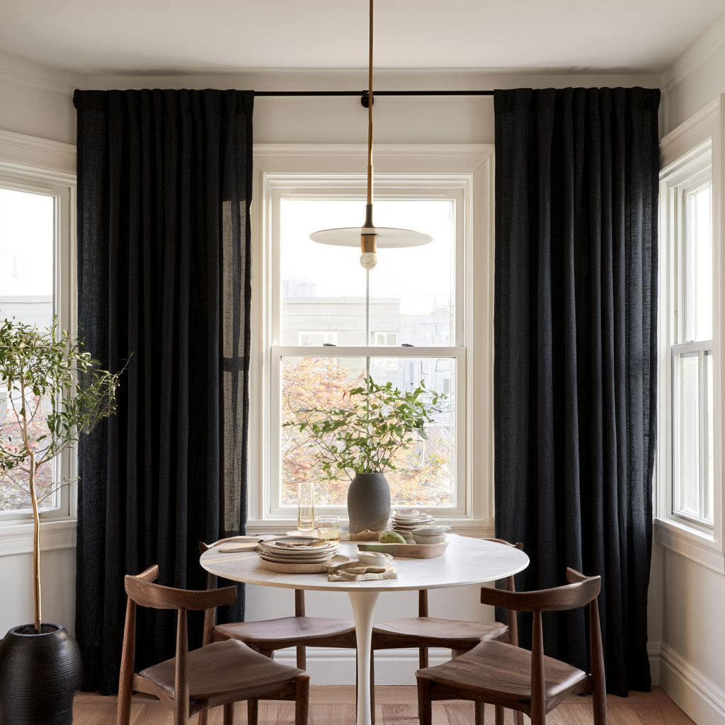 Floor-length black curtains frame a bright dining room window behind a round white table with wooden chairs, stacked dishes, and a vase of leafy branches