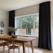 Floor-length black curtains frame a wide dining room window overlooking trees, beside a wooden table and chairs with a vase of flowers and a bowl of fruit