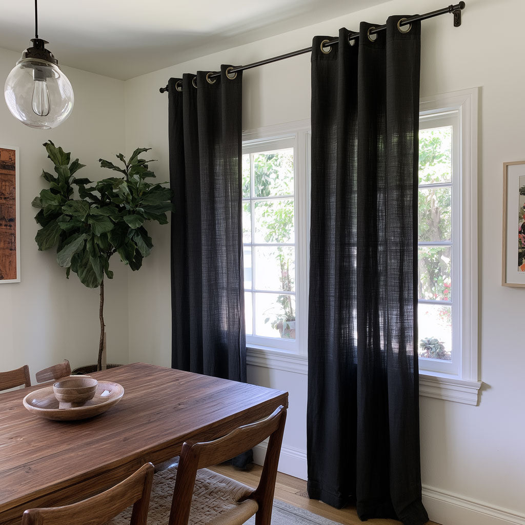Floor-length black eyelet curtains hang beside two dining room windows, with a wooden table, woven-seat chairs, a tall leafy plant, and a glass pendant light