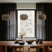 Floor-length black curtains frame a large dining room window behind a wooden table with ceramic dishes, wooden chairs, and three cream pendant lights