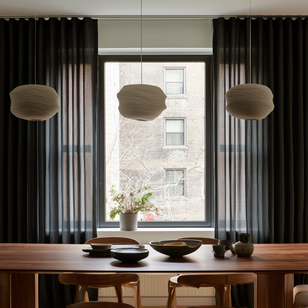 Floor-length black curtains frame a large dining room window behind a wooden table with ceramic dishes, wooden chairs, and three cream pendant lights