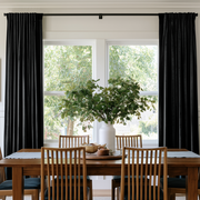 Bright dining room with black floor-length curtains, warm wooden furniture, and fresh greenery