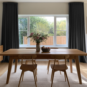 Floor-length charcoal gray curtains frame a wide window behind a wooden dining table with matching chairs, a vase of flowers, and a bowl of fruit