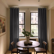 Floor-length charcoal gray eyelet curtains frame two narrow windows behind a round wooden dining table with wooden chairs, stacked bowls, and a vase of leafy branches