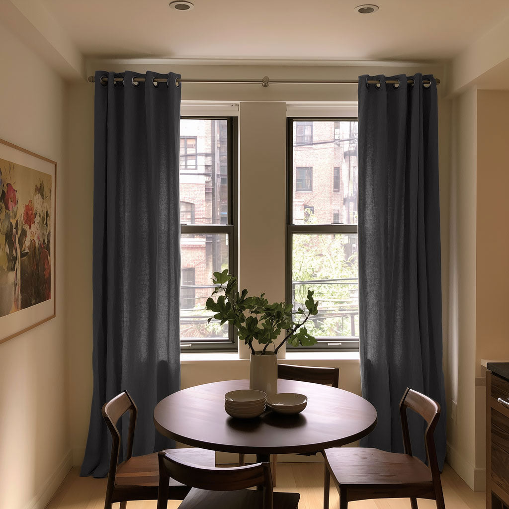 Floor-length charcoal gray eyelet curtains frame two narrow windows behind a round wooden dining table with wooden chairs, stacked bowls, and a vase of leafy branches