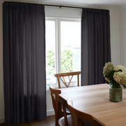 Floor-length charcoal gray pinch-pleat curtains frame a bright window beside a wooden dining table and chairs, with pale hydrangeas in a green glass vase
