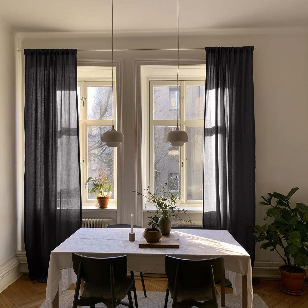 Charcoal gray curtains frame two tall windows in a sunlit dining room with a white-covered table, black chairs, two pendant lights, and potted plants