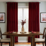 A stylish dining room with rich burgundy curtains, a dark wooden table, upholstered chairs, coordinated red napkins, and a tall decorative vase with dried branches