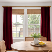 A bright dining room with rich burgundy curtains, woven Roman shades, an oval wooden table, cane-back chairs, and a simple centerpiece with greenery and fresh lemons