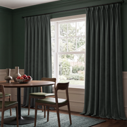 A moody dining room with deep green walls, charcoal-gray curtains, a round wooden table, mid-century chairs with green cushions, sculptural vases, and a bowl of red apples