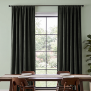 A minimalist dining room with floor-length charcoal curtains, a large black-framed window overlooking a garden, a long wooden table, and warm brown chairs