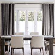 A minimalist dining room with dark gray pleated curtains, large white French doors, a dark wooden table, cream upholstered chairs, and a simple light table runner
