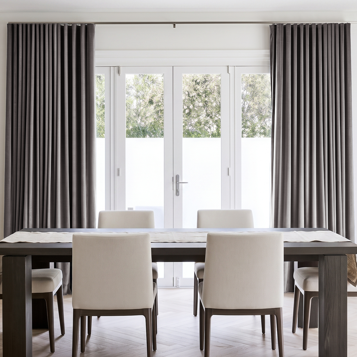 A minimalist dining room with dark gray pleated curtains, large white French doors, a dark wooden table, cream upholstered chairs, and a simple light table runner