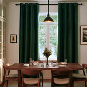 A stylish dining room with deep forest-green curtains, a long wooden table, matching chairs, elegant tableware, a floral centerpiece, and a black pendant light
