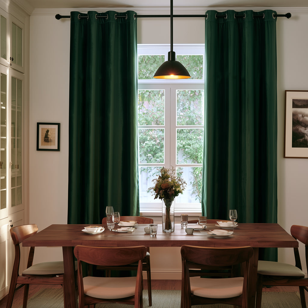 A stylish dining room with deep forest-green curtains, a long wooden table, matching chairs, elegant tableware, a floral centerpiece, and a black pendant light