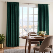 A bright sophisticated dining room with deep emerald curtains, a round wooden table, taupe upholstered chairs, elegant place settings, and a low floral centerpiece