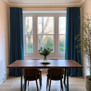 Floor-length teal blue curtains frame a wide window behind a wooden dining table and chairs, with leafy branches in a glass vase and a tall potted tree nearby