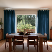 Floor-length teal blue eyelet curtains frame a large window overlooking trees behind a wooden dining table with matching chairs, dark dishes, and a small vase of greenery