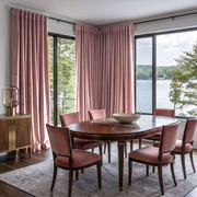 A modern lakeside dining room with soft blush-pink curtains, panoramic windows, an oval dark-wood table, matching pink upholstered chairs, and a minimalist centerpiece