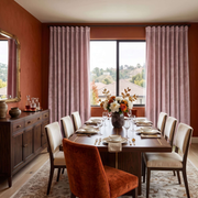 An elegant dining room with terracotta walls, soft blush-pink curtains, a dark wooden table, cream upholstered chairs, formal place settings, and a rich autumn floral centerpiece