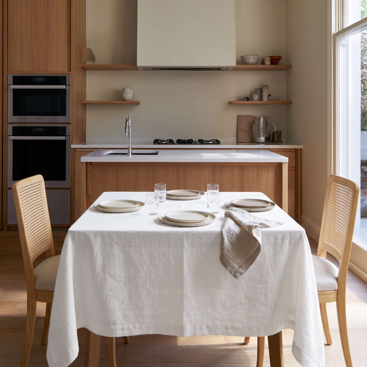 Off-White Square Linen Tablecloth in a Minimalist Kitchen with Cane Dining Chairs and Warm Wood Cabinetry