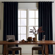 A cozy dining room with floor-length black curtains, a wooden table, upholstered gray chairs, dark ceramic tableware, and a decorative vase with burgundy branches