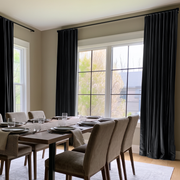 A bright contemporary dining room with floor-length black curtains, large corner windows, a dark wooden table, upholstered taupe chairs, and neatly arranged tableware