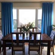 A cozy dining room with vibrant blue curtains, a dark wooden table, mixed upholstered and wooden chairs, and a simple leafy centerpiece in front of a bright window