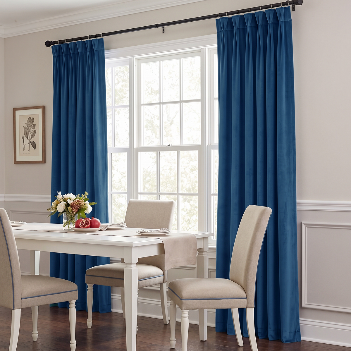 A bright elegant dining room with rich blue curtains, a white wooden table, cream upholstered chairs, fresh flowers, fruit, and abundant natural light