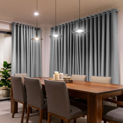 Floor-length gray eyelet curtains cover dining room windows behind a wooden table with beige upholstered chairs, a tray of candles, and three glowing pendant lights