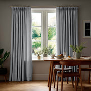 Floor-length gray pinch-pleat curtains hang from a brass rod around a dining room window beside a wooden table set with dishes, glasses, and leafy plants