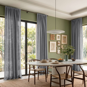 Floor-length gray curtains frame large windows in a sage green dining room with a white oval table, woven-seat wooden chairs, and a white pendant light