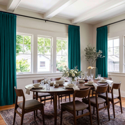 A spacious dining room with rich teal curtains, large corner windows, a dark wooden table, classic chairs, elegant tableware, and a fresh floral centerpiece